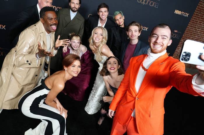 Group of people dressed stylishly, posing for a selfie at a red carpet event. Everyone is smiling and facing the camera