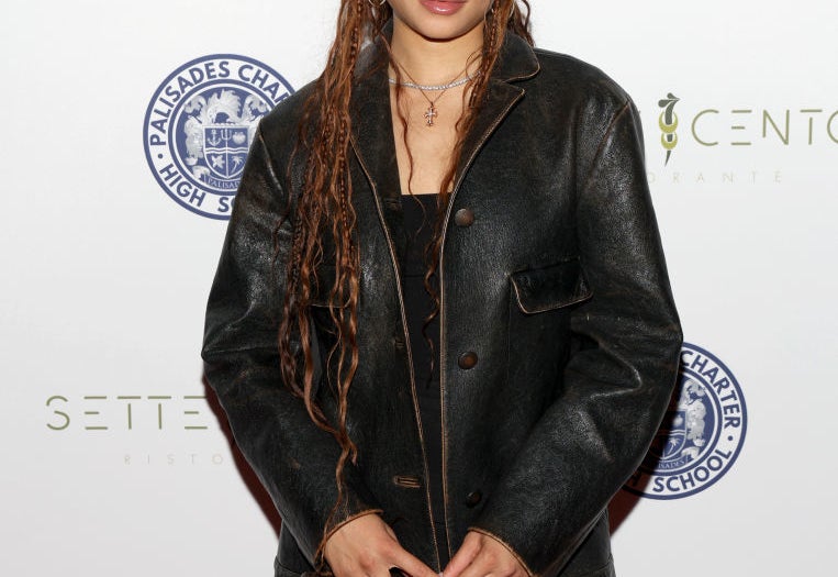 Person in a leather jacket with long braided hair on a red carpet at an event, posing in front of a backdrop with logos
