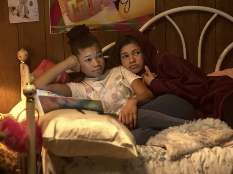 Two young women relax on a bed, one in a hoodie. Both appear focused while looking at an unseen laptop. Cozy bedroom setting with posters on the wall