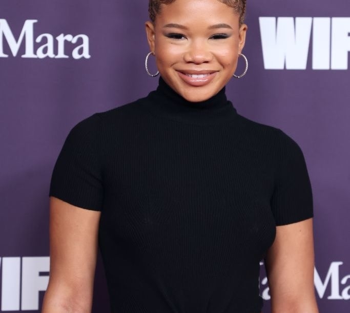 Person in a black turtleneck crop top with unique braided hairstyle poses at a celebrity event backdrop