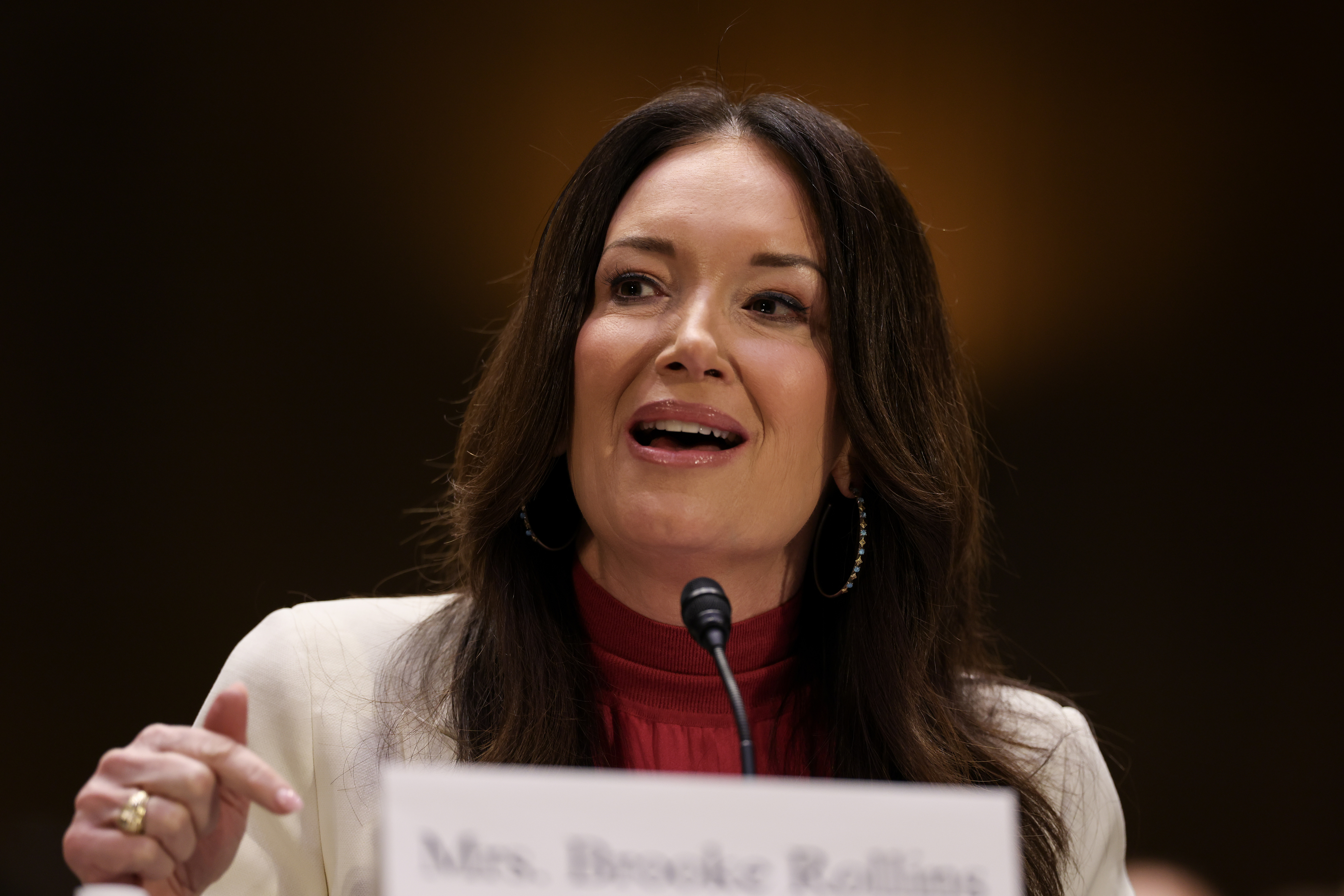 Brooke Rollins in a white blazer speaks at a podium with a microphone during a formal event