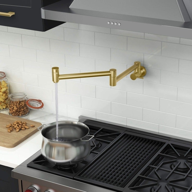 A brass pot filler faucet extends over a pot on a stove, with kitchen items like jars and cutting boards in the background