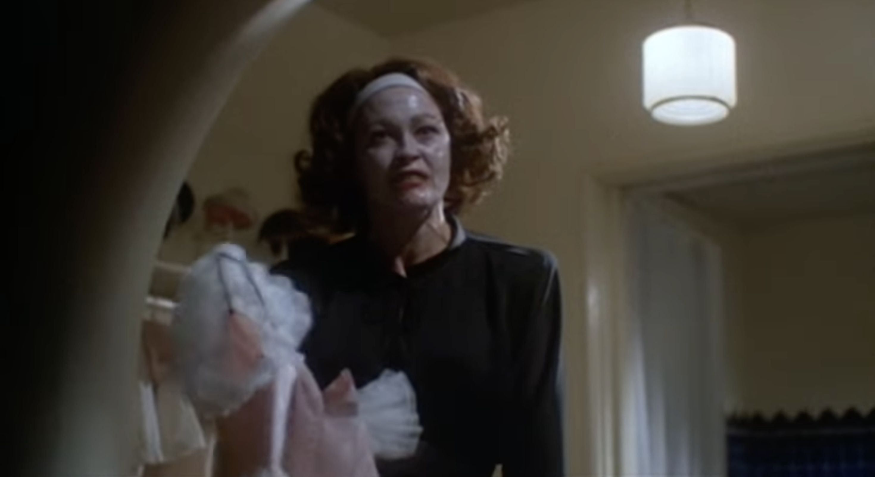 Person with a white facial mask, wearing a black outfit and headband, holds a ruffled garment, standing in a dimly lit room