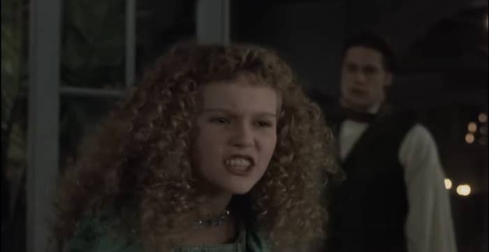 A young woman with curly hair looks frustrated, while a man in a vest stands behind her in a dimly lit room