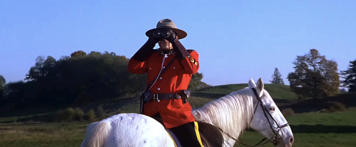 A Mountie in traditional uniform on a white horse uses binoculars to survey a grassy landscape