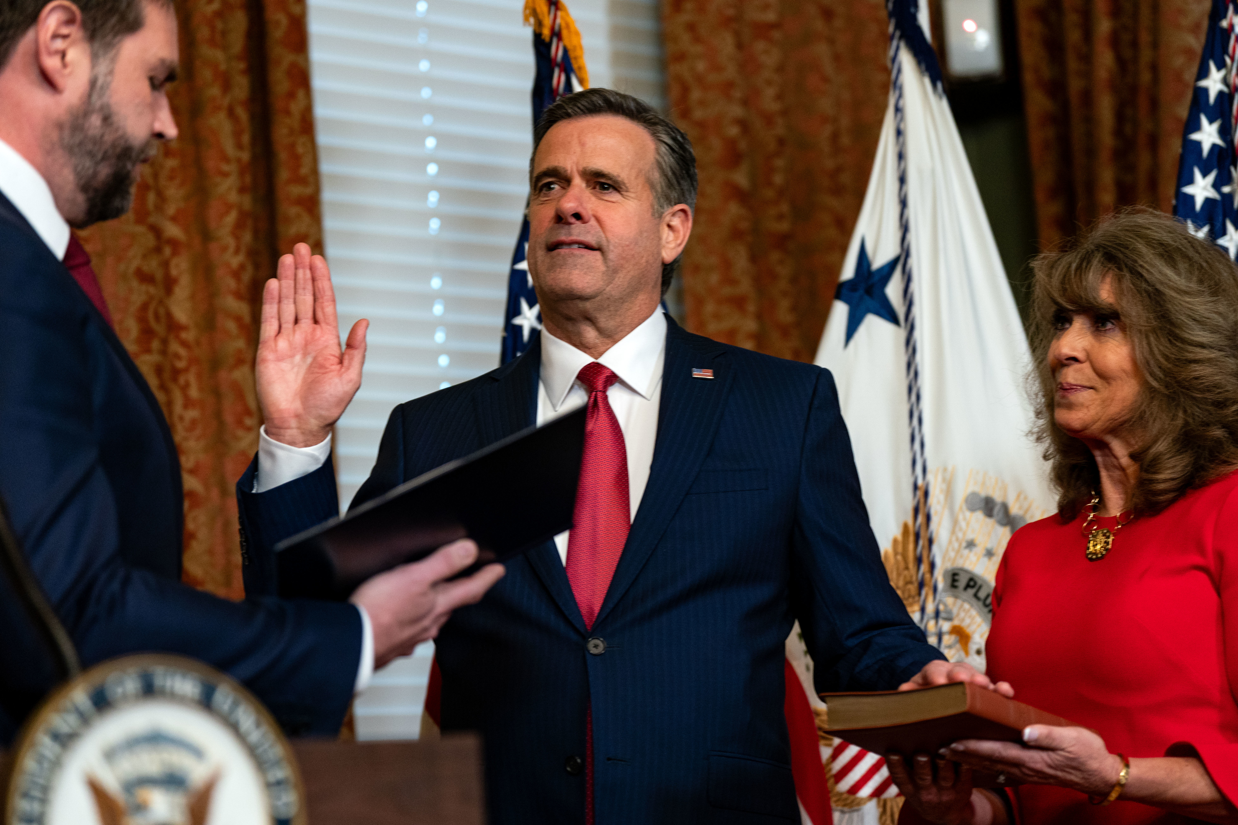 John Ratcliffe being sworn in, raising his right hand. A woman holds a Bible for the ceremony. U.S. flags are in the background