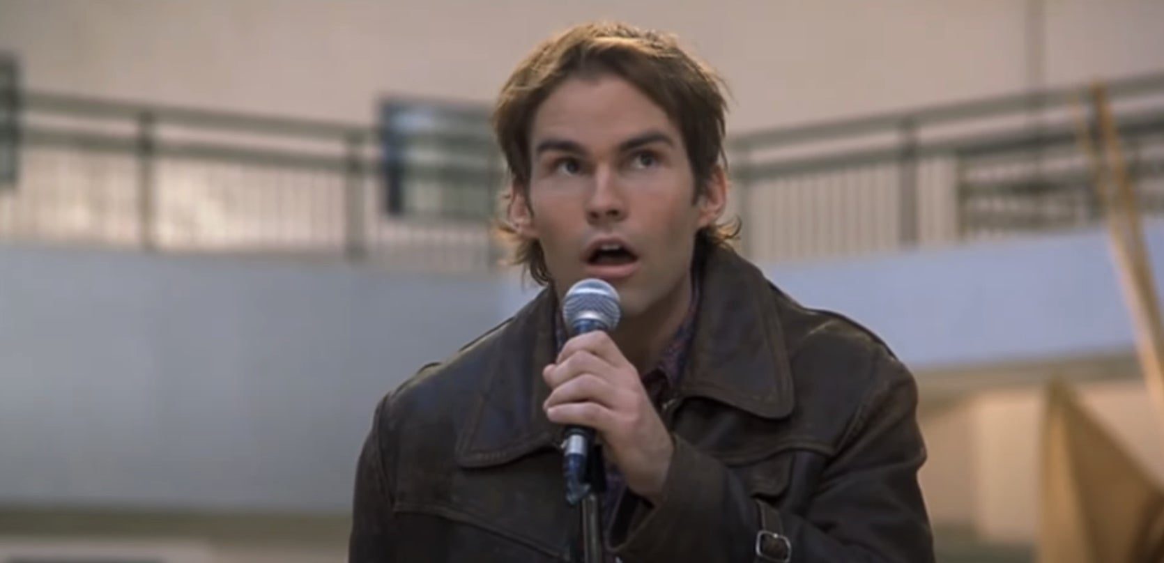 A person in a leather jacket holds a microphone and appears surprised or in mid-speech in an indoor setting