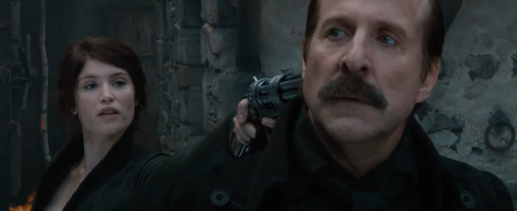 A woman points a revolver at a man's head in an intense scene, possibly from a film. They appear in a dim, gritty setting