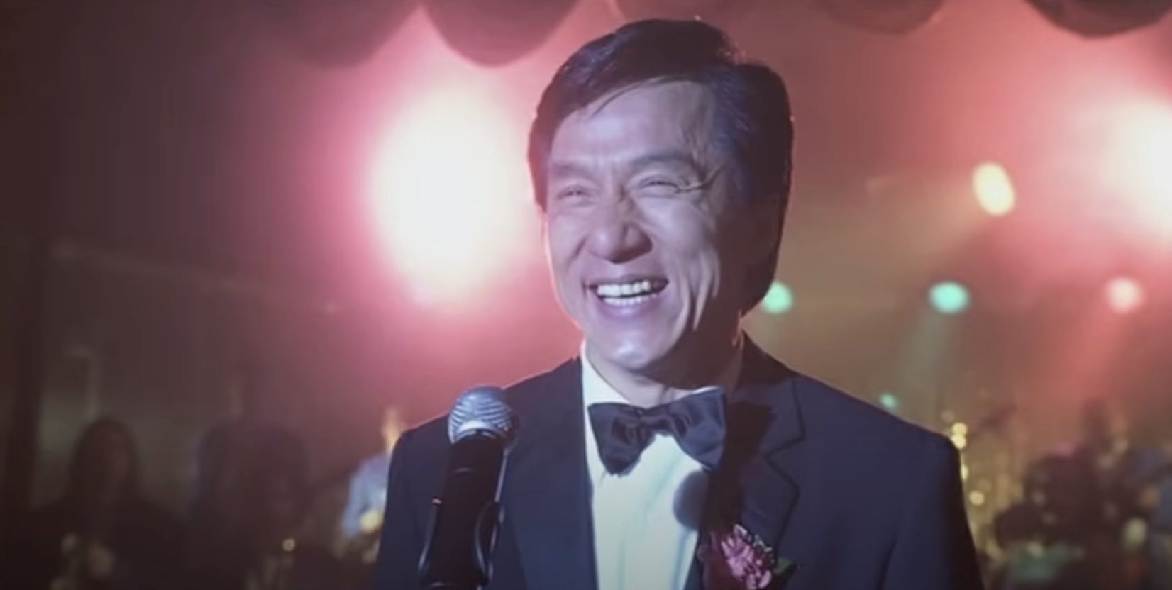 A person in a tuxedo is smiling broadly while standing in front of a microphone on stage with bright lights in the background