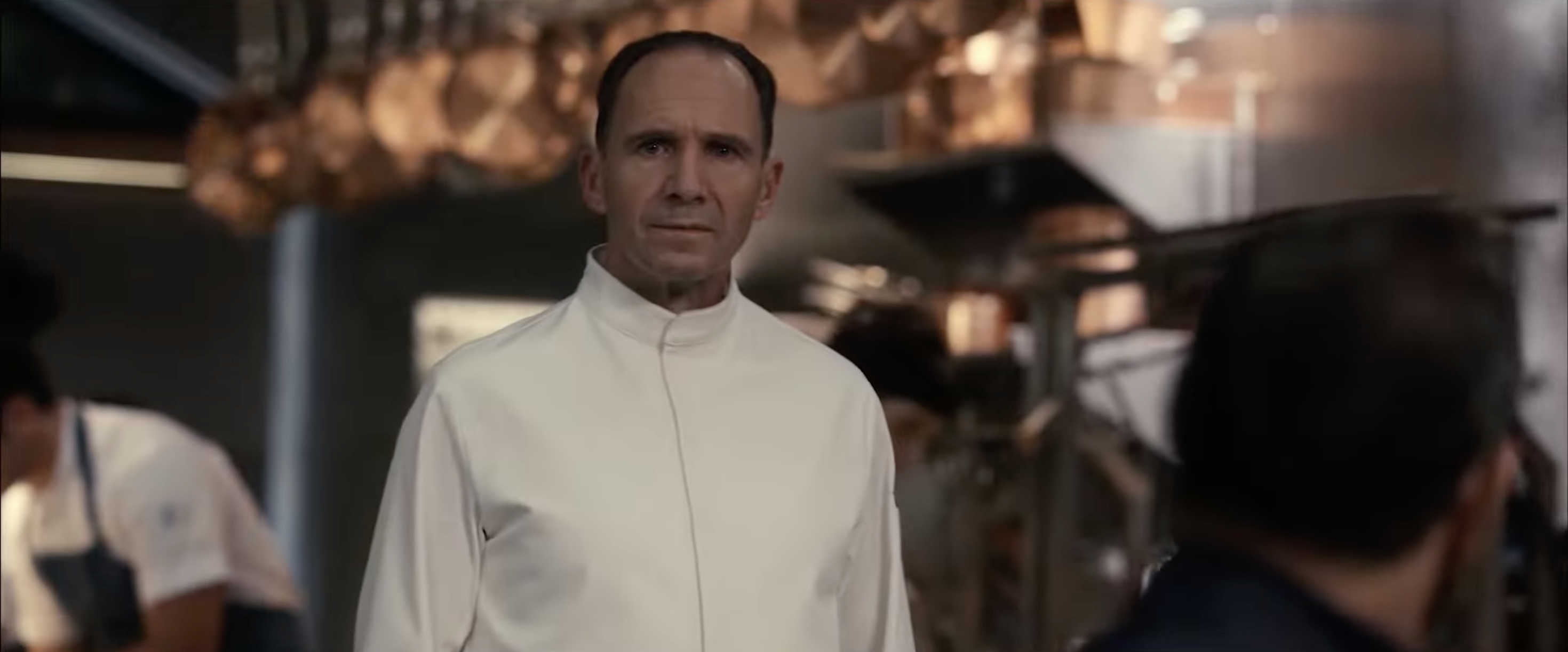 Chef in a pristine kitchen wearing a white chef's coat, surrounded by copper pans and busy staff