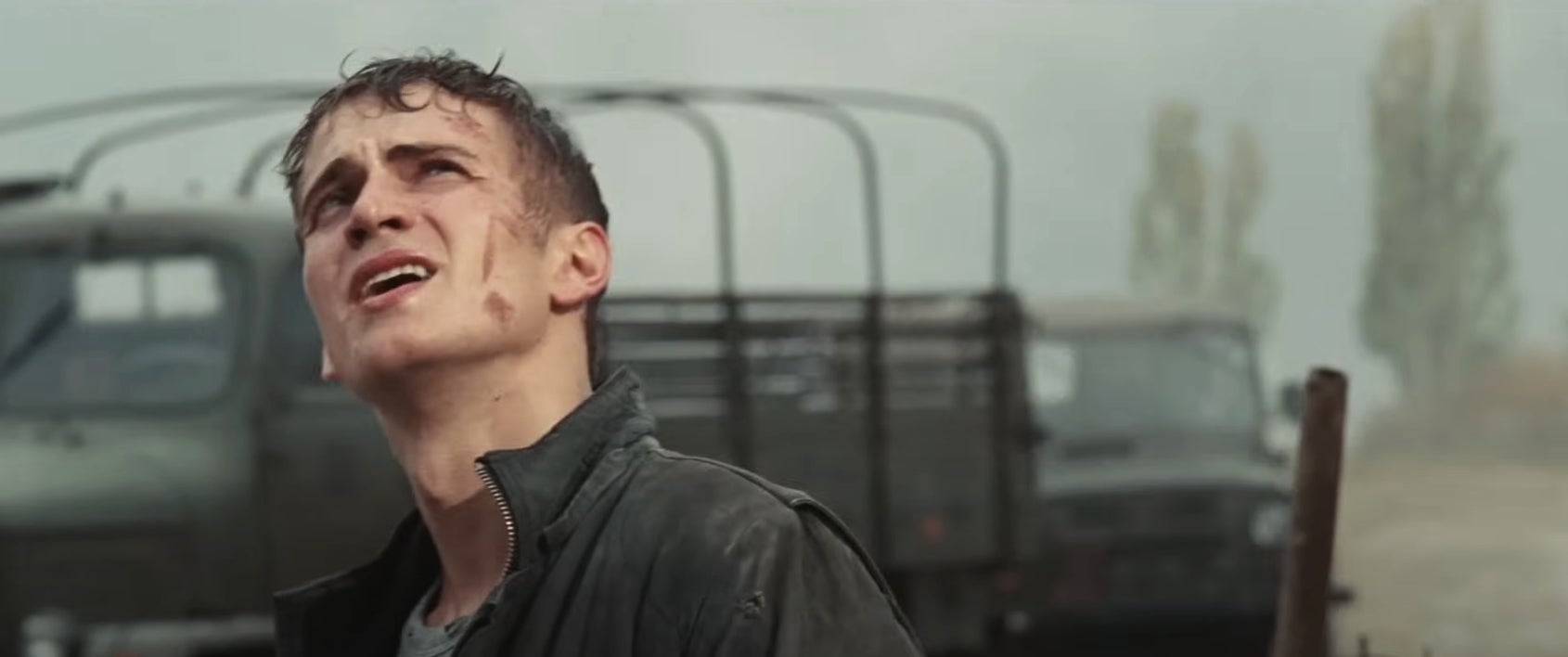 A young man with a dirt-streaked face looks upward, standing in front of military trucks in a bleak outdoor setting