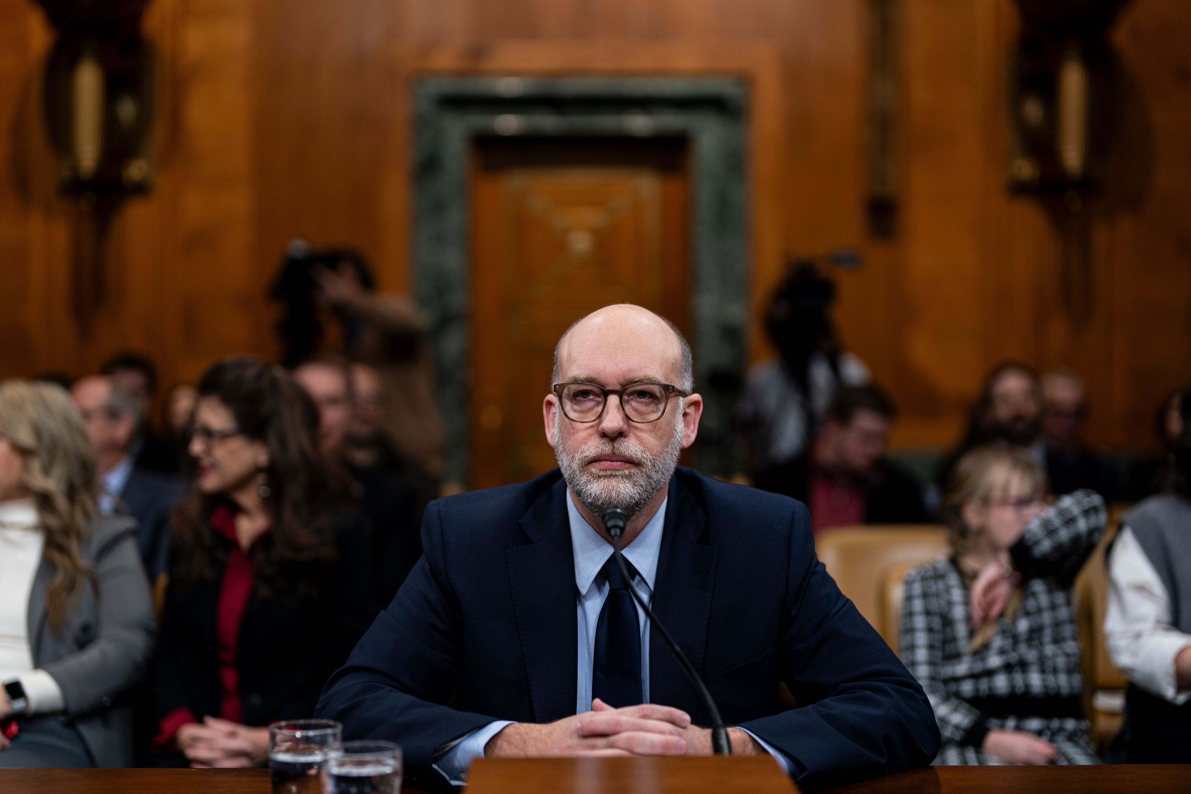 Russell Vought sits at a table during a formal hearing, with people seated in the background