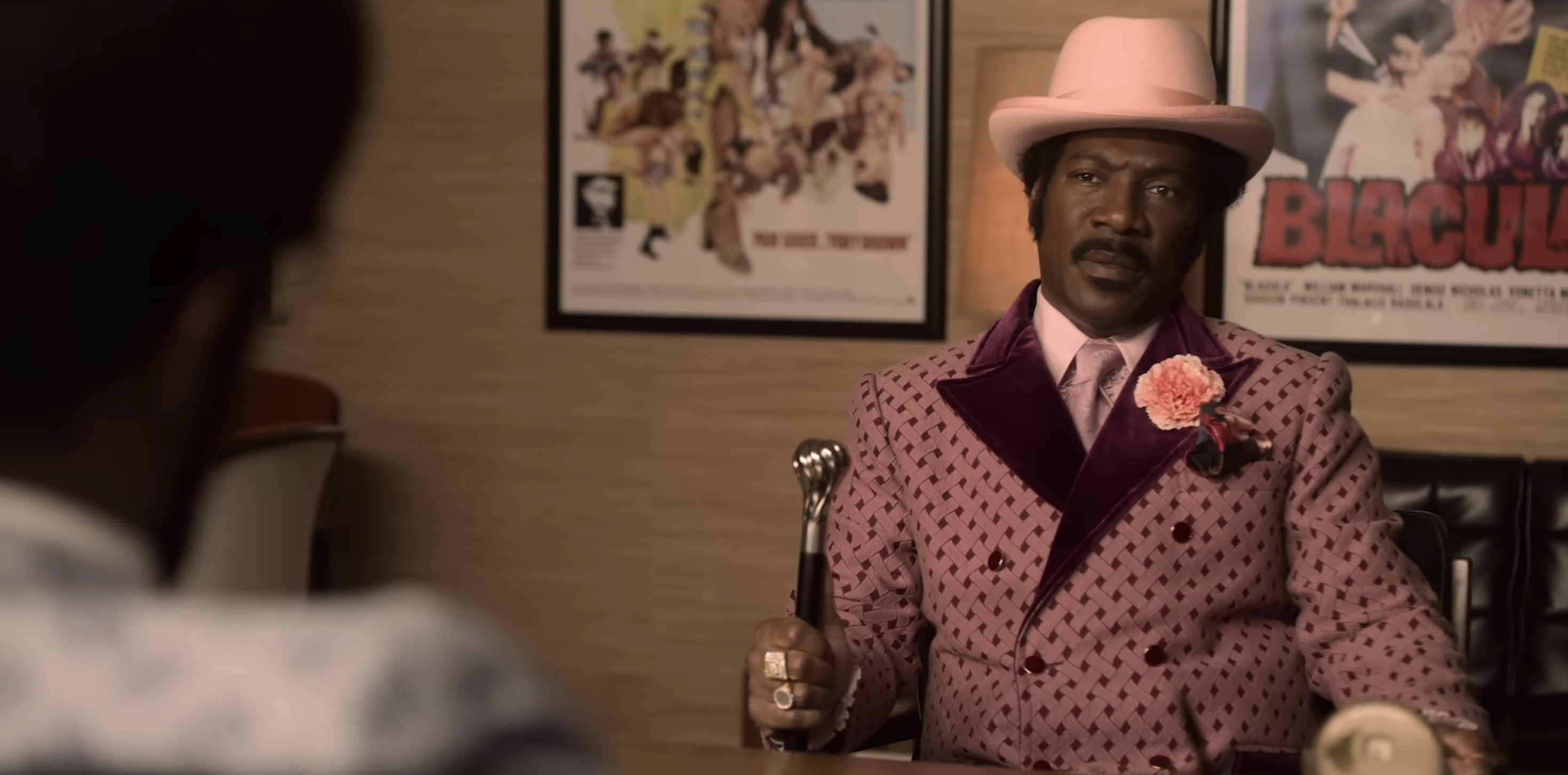 Man in vintage-style suit with a polka dot pattern and wide-brimmed hat sits at a table holding a microphone. Movie posters are on the wall behind him