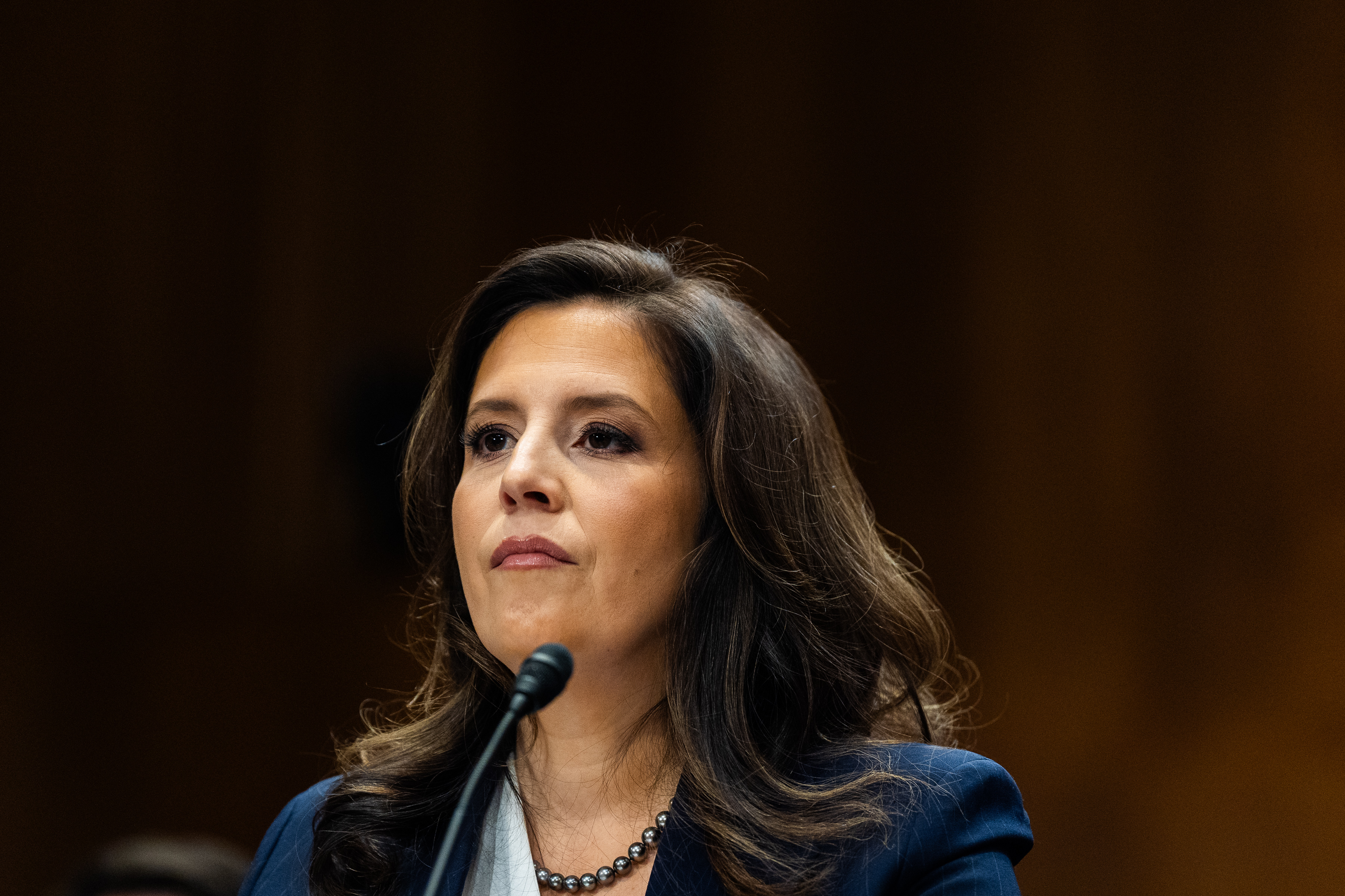 Elise M. Stefanik speaks into a microphone, wearing a business suit and necklace, against a blurred background