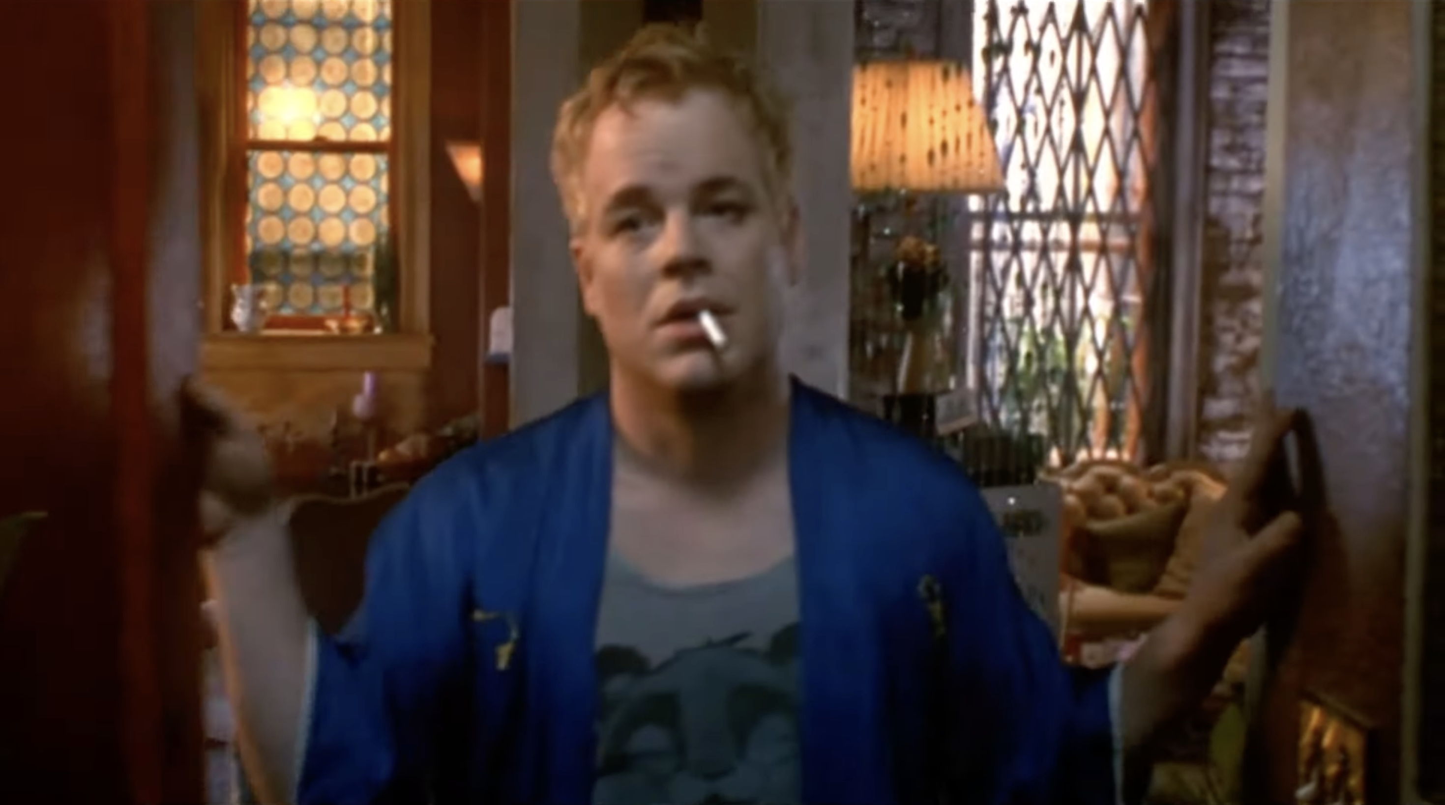 A person in a film scene stands in a doorway, wearing a casual robe and t-shirt, holding cigarettes in their mouth. The background shows a cozy interior