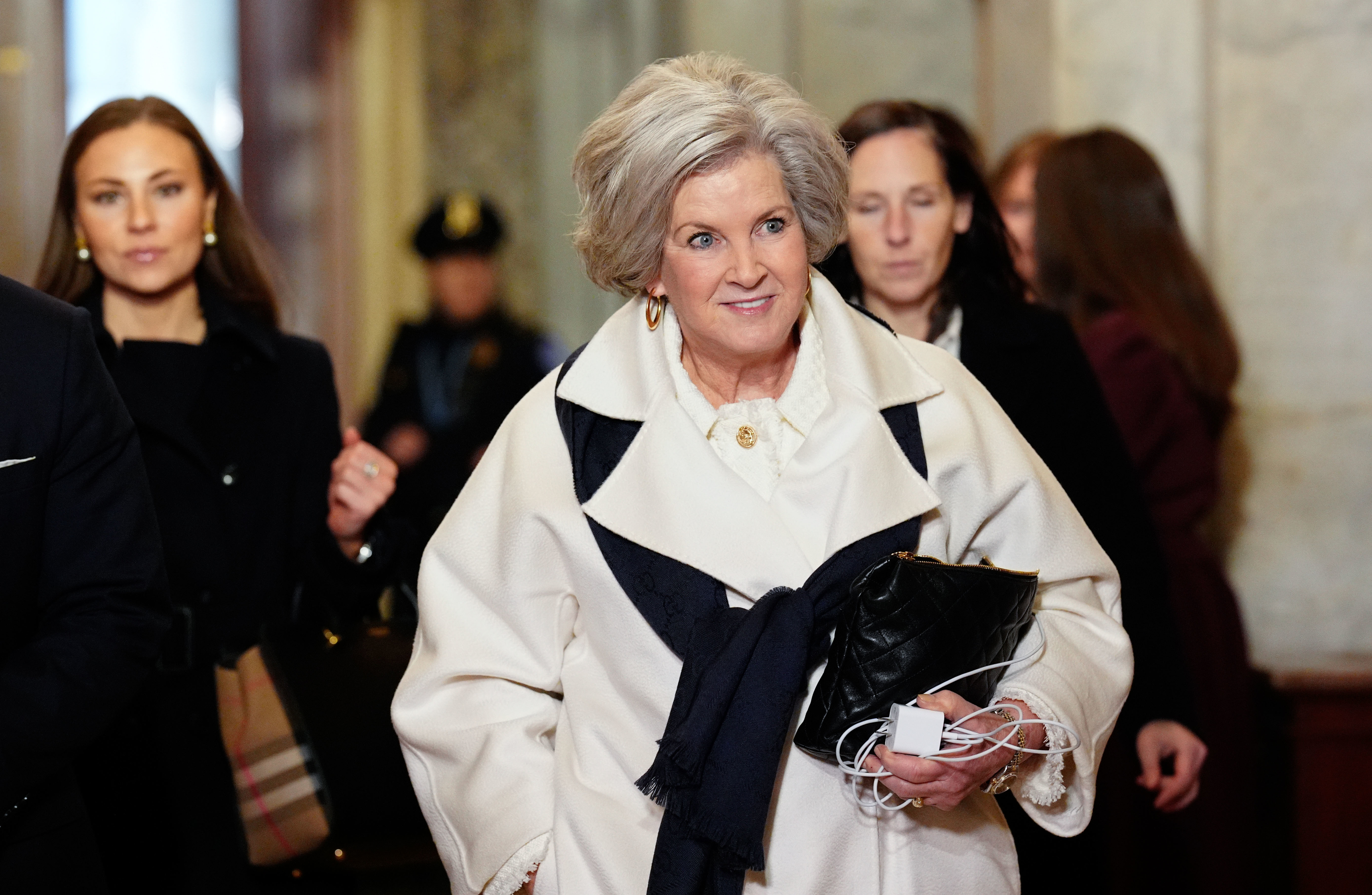 Susie Wiles in a stylish coat with a scarf, holding a black clutch and headphones, walking inside with a group of people