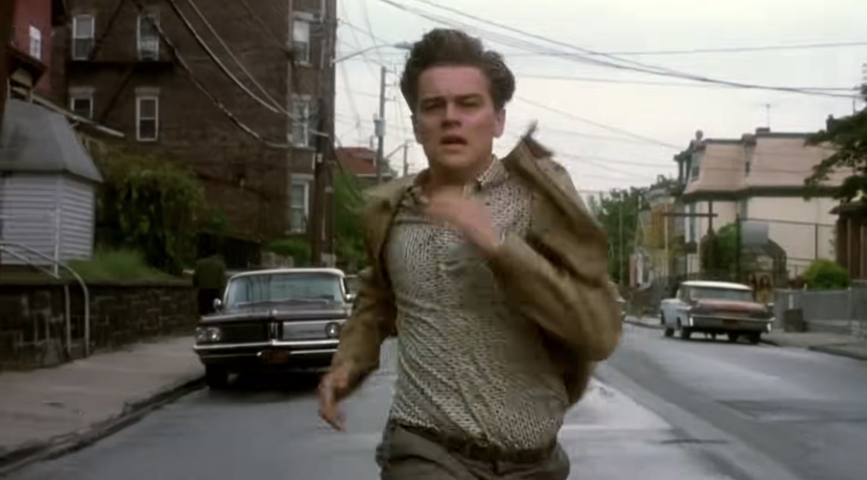 Person in a casual jacket and patterned shirt running on an empty street, with classic cars in the background