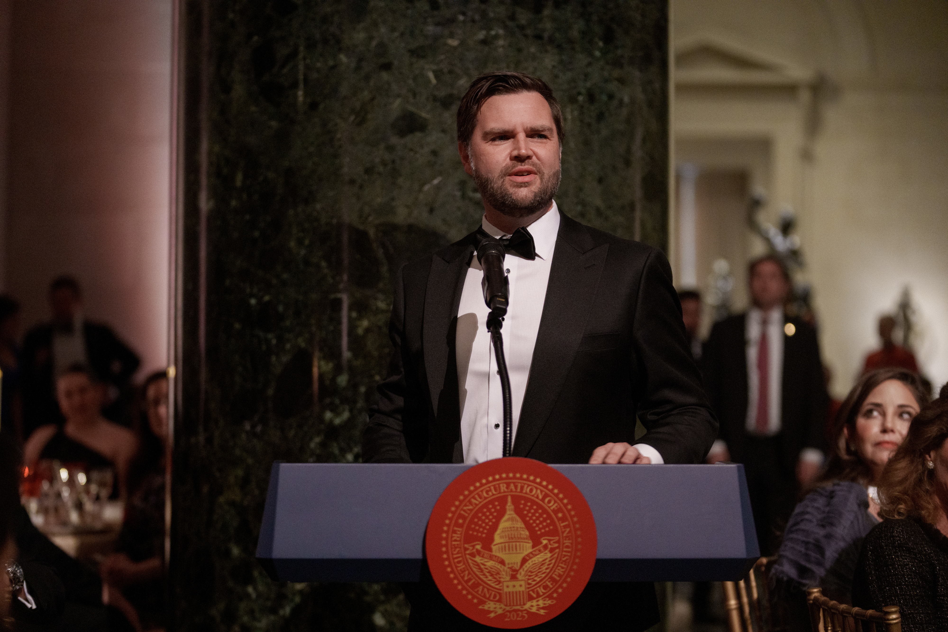 JD Vance in a tuxedo speaks at a podium with a seal, in a formal event setting. Audience members are seated at tables in the background