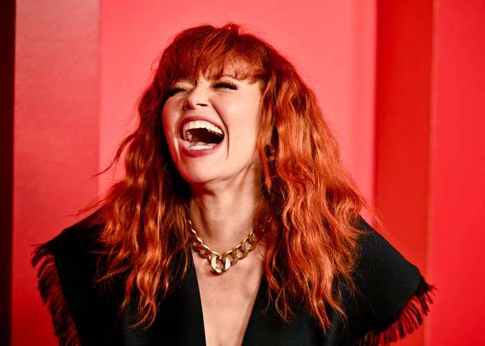Person with long wavy hair, wearing a statement necklace, laughing joyfully in front of a red backdrop