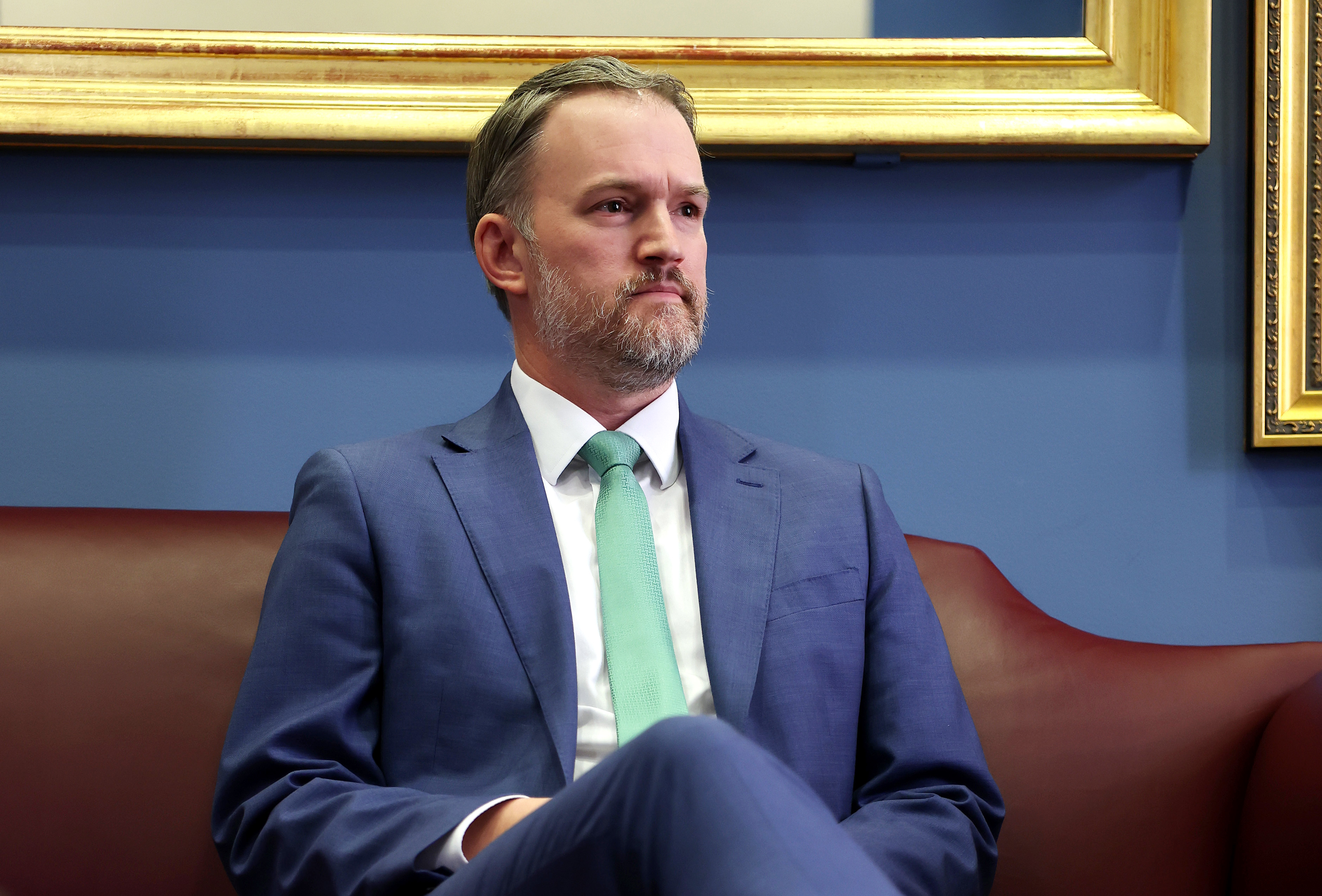 Jamieson Greer, sitting on a brown couch, looking to the side. Background features a gold-framed picture