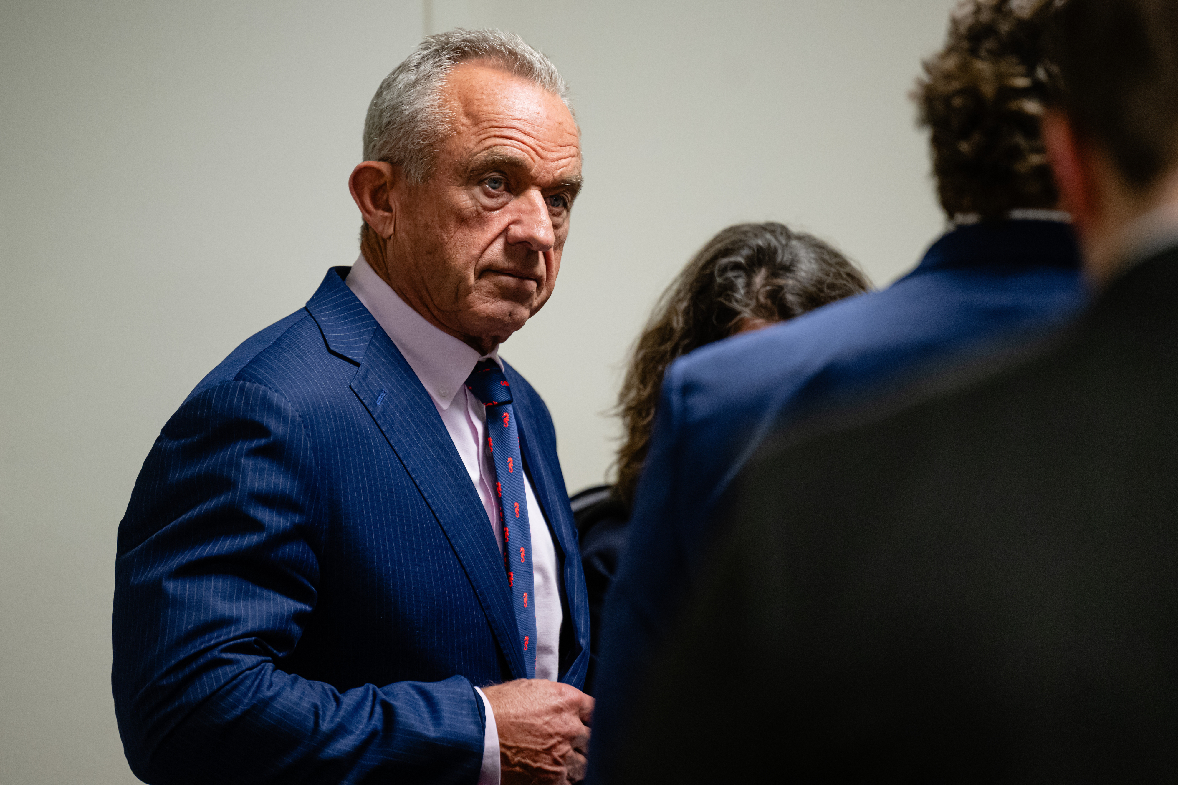 RFK Jr. in a suit stands among a group, appearing focused and attentive in a formal setting