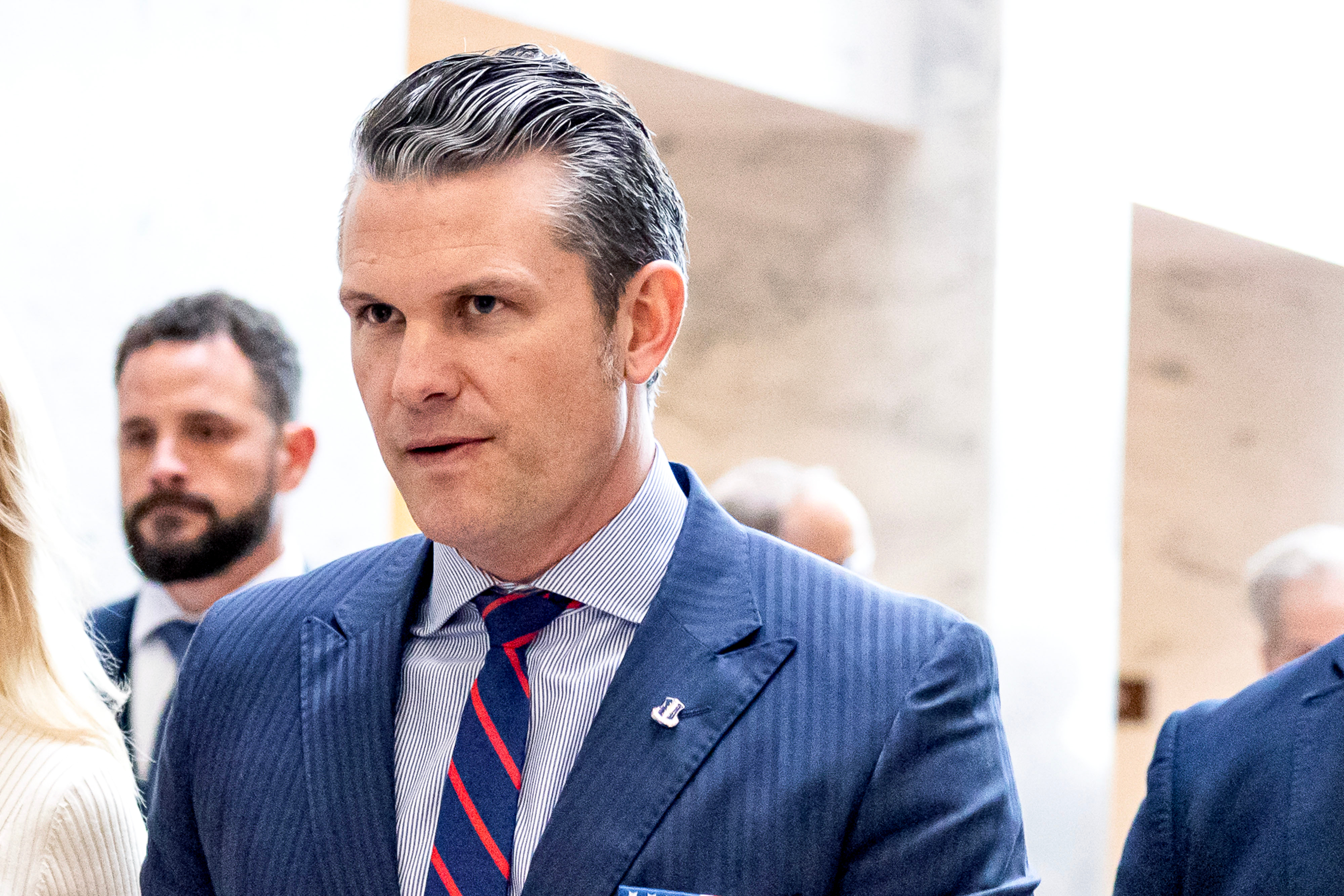 Pete Hegseth in a pinstripe suit and blue shirt with a striped tie appears walking indoors, accompanied by others