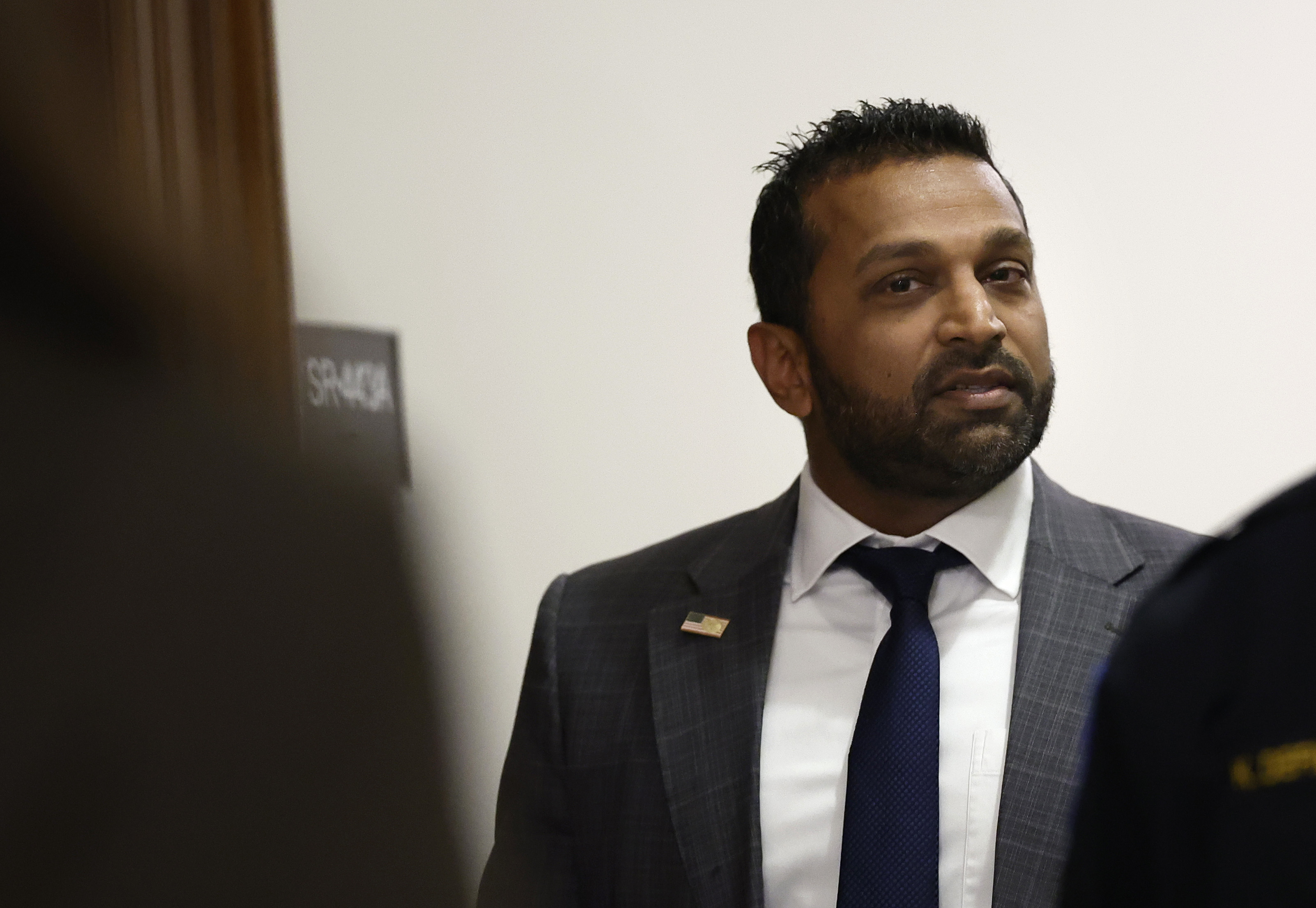A man in a suit and tie stands in a hallway, looking to the side with a thoughtful expression