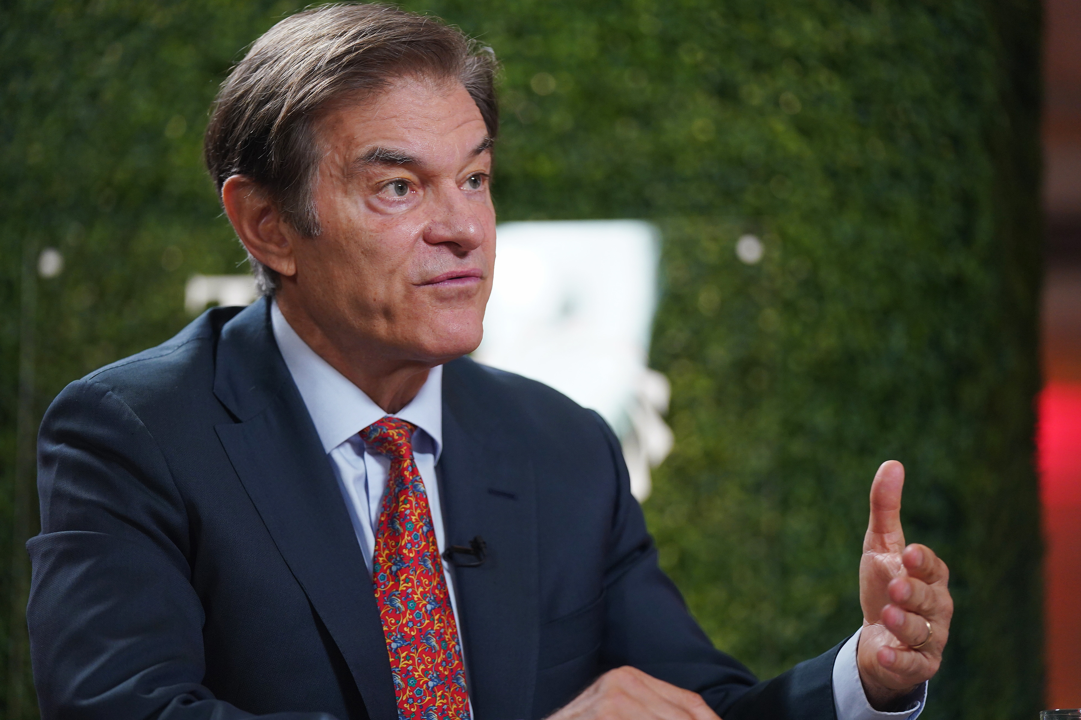 Dr. Oz in a suit gestures while speaking in an outdoor setting with greenery in the background