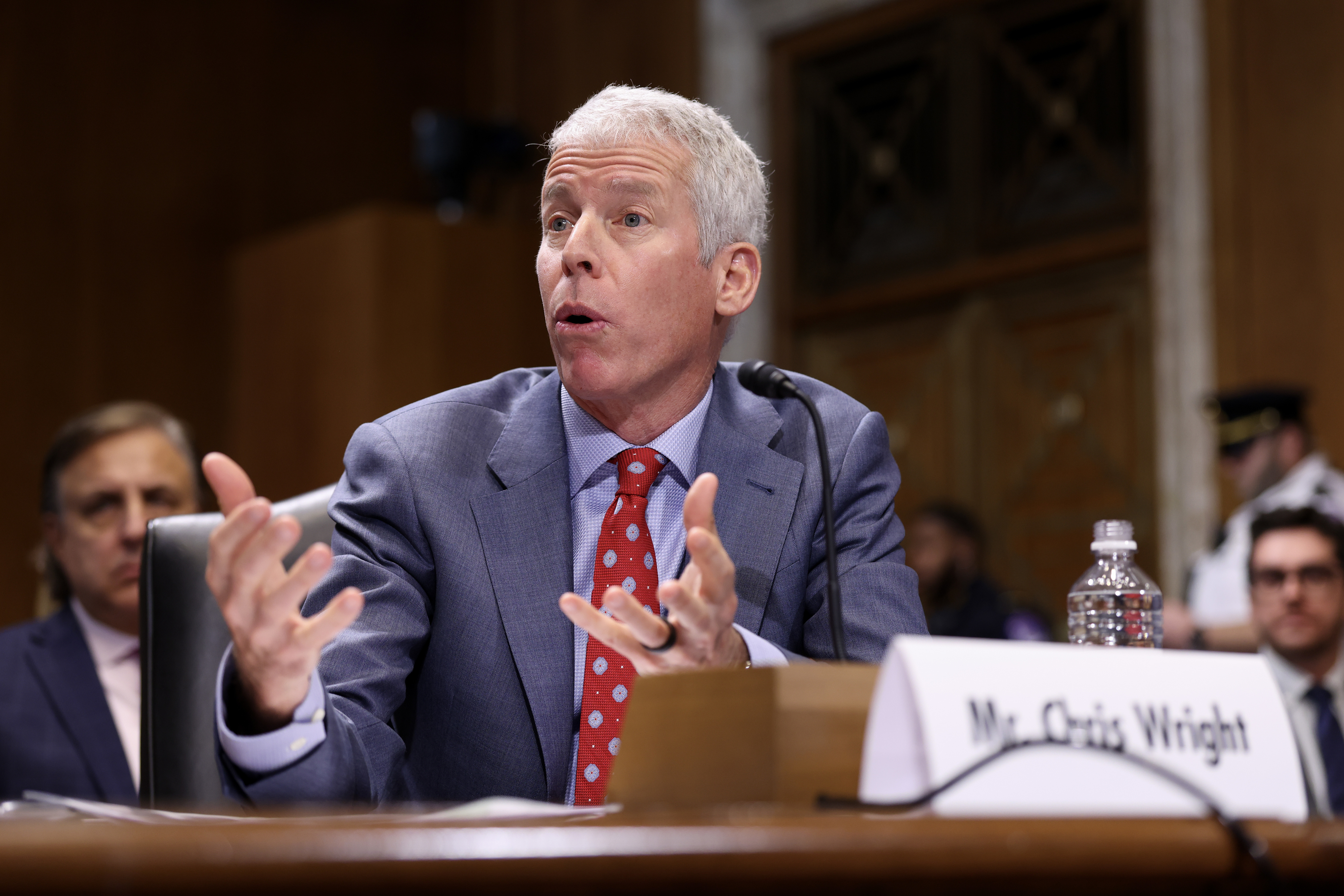 Man in a suit gesturing while speaking at a panel with a microphone. Nameplate reads "Mr. Chris Wright." Others are seated in the background