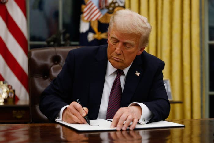 A person in a suit is sitting at a desk in an ornate office, focused on signing a document