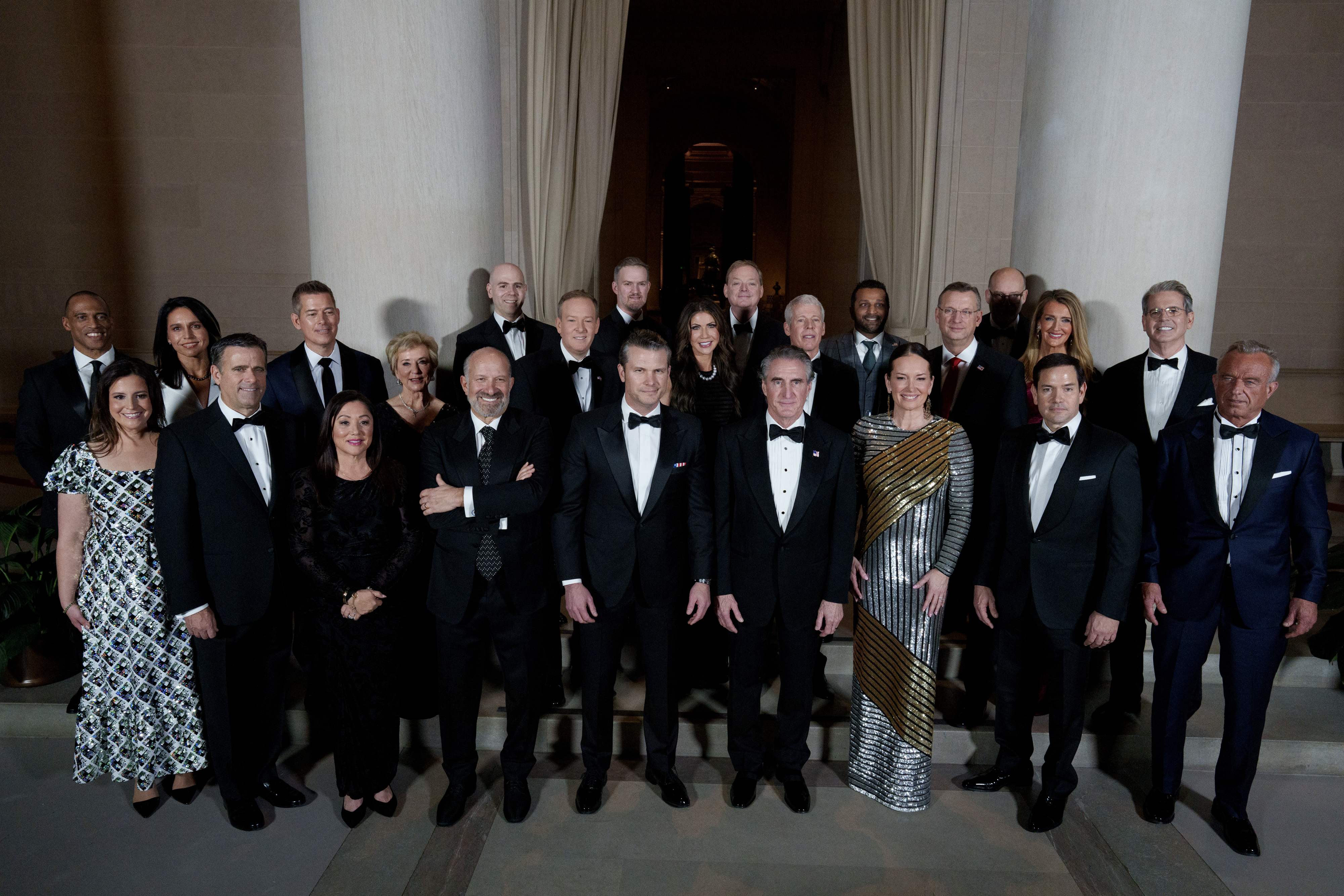 Group of individuals dressed in formal evening attire, including tuxedos and gowns, posed in an elegant setting with tall columns