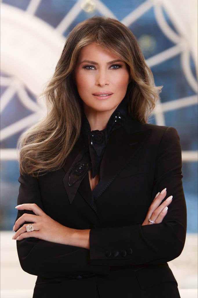 Melania in a formal suit with arms crossed, standing indoors with a blurred background