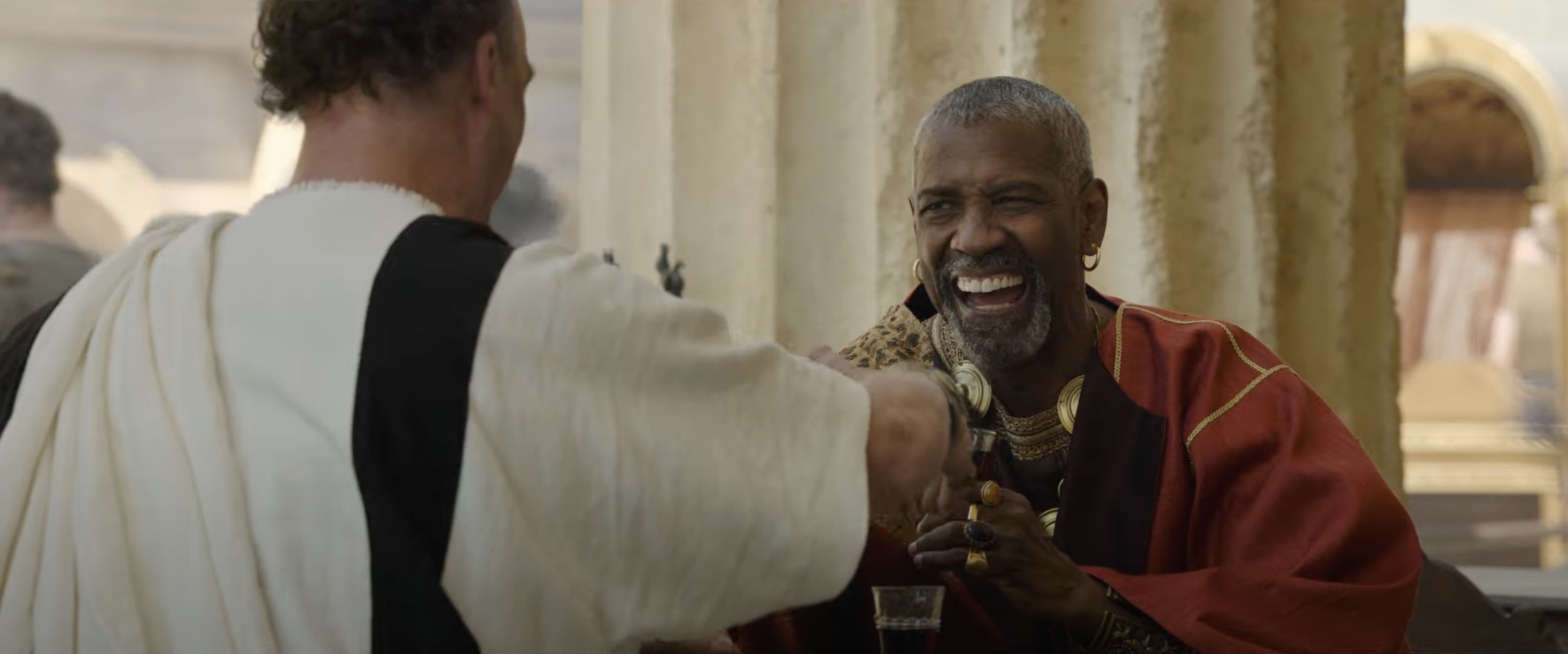 Two people in ancient Roman-style attire share a moment. One, draped in a red toga, smiles widely while holding a drink
