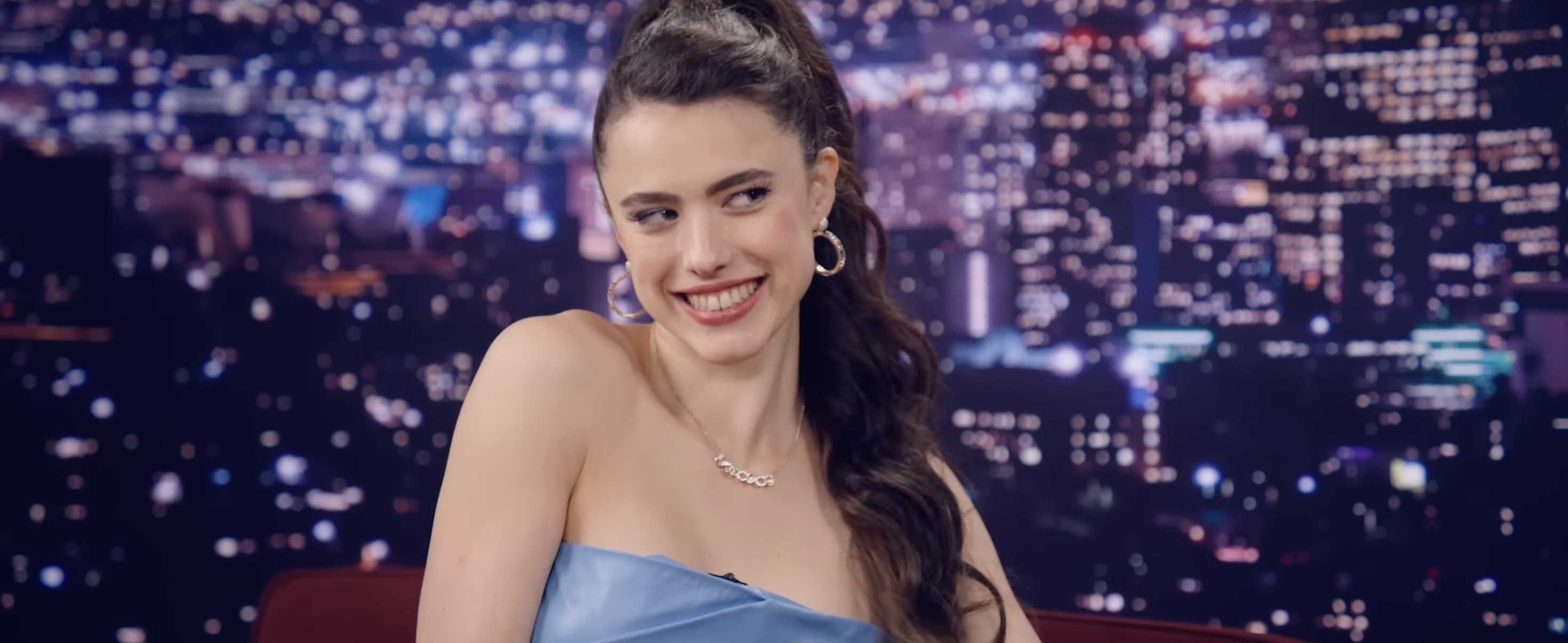 Person in strapless dress smiling on a talk show set with a cityscape background