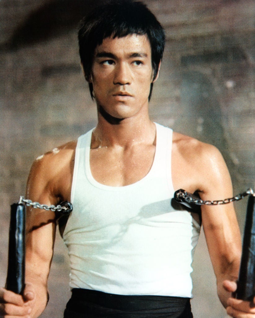Bruce in a tank top holds weapons, appearing focused and ready for action