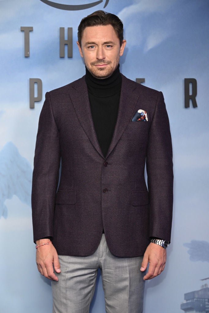 JJ in a dark blazer, turtleneck, and light trousers stands against a backdrop at an event