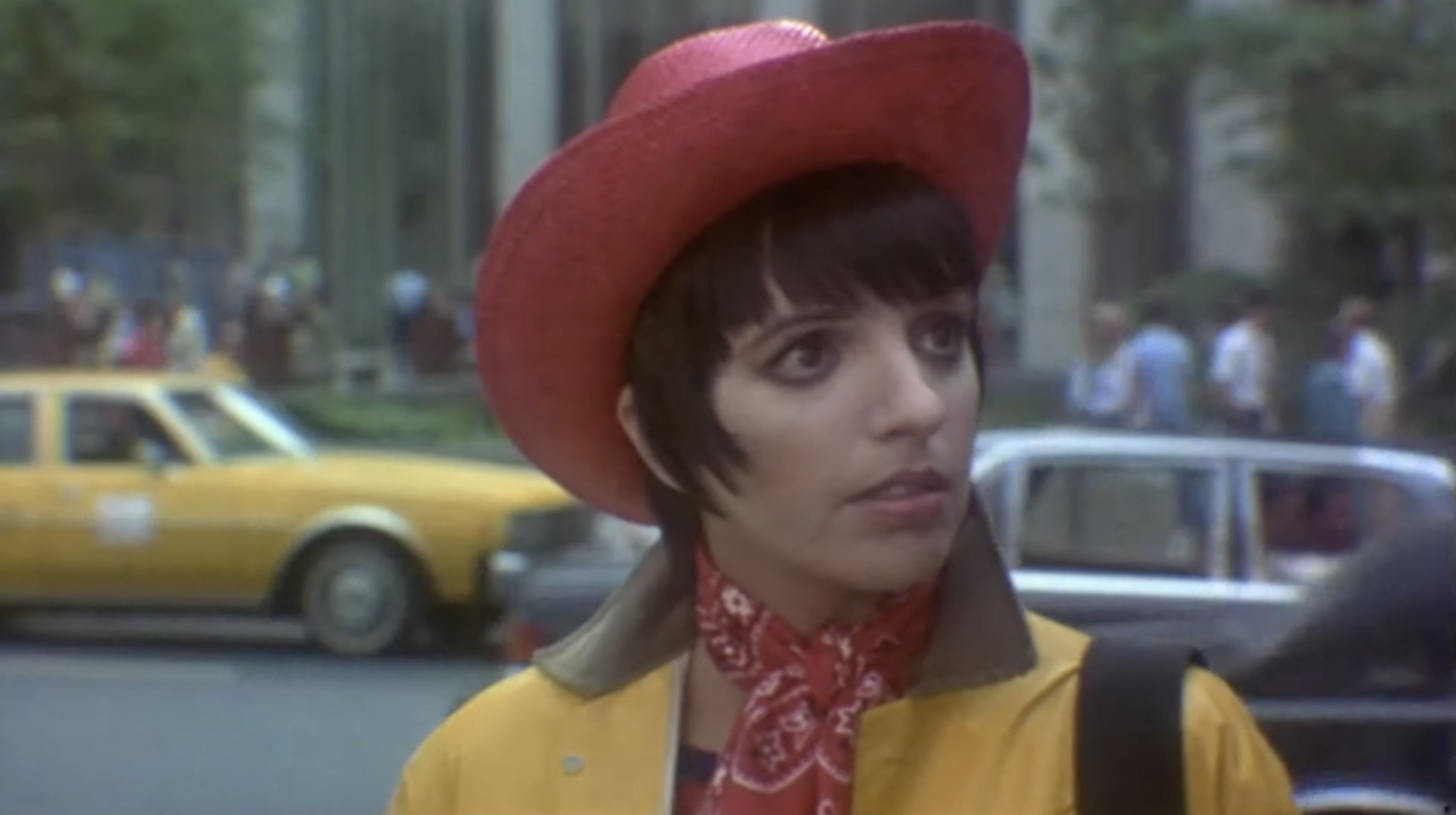 Person wearing a red cowboy hat and bandana, standing on a city street with taxis and buildings in the background