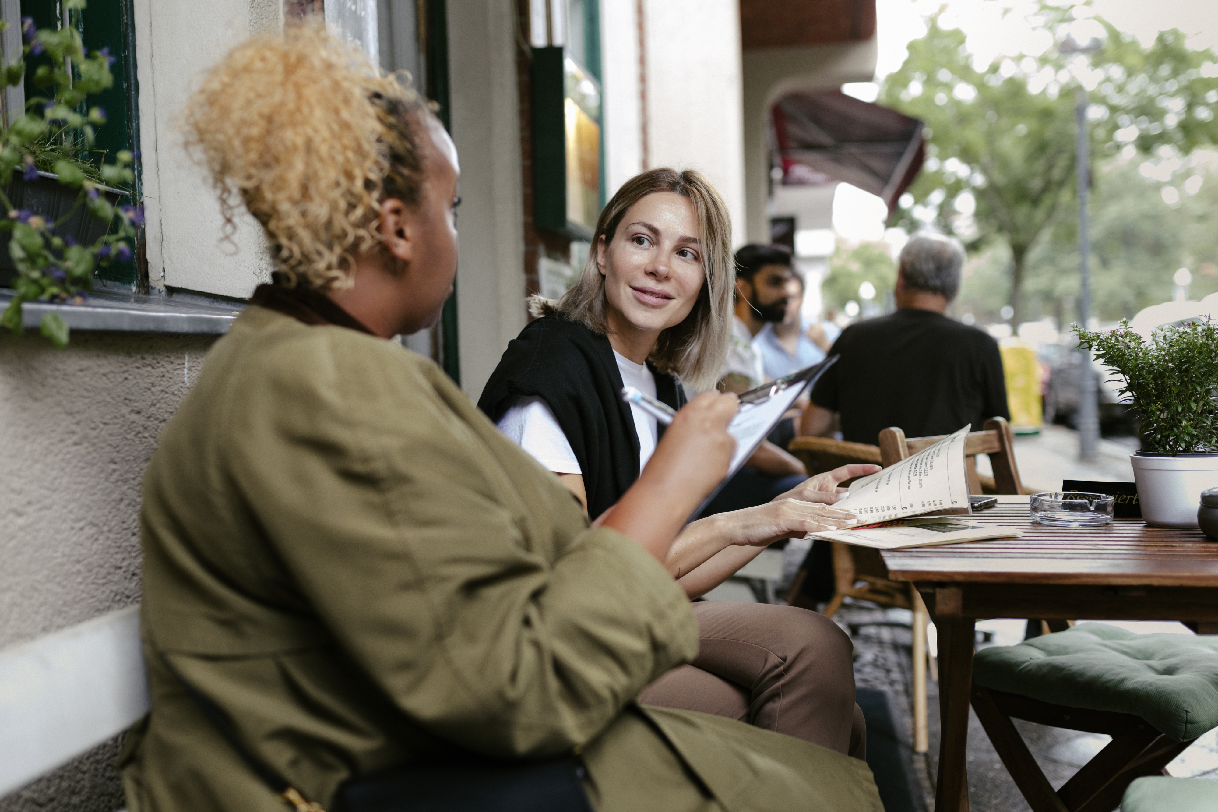 To personer sitter utendørs på en kafé. Den ene holder en meny og snakker med den andre. Mennesker og bord er synlige i bakgrunnen