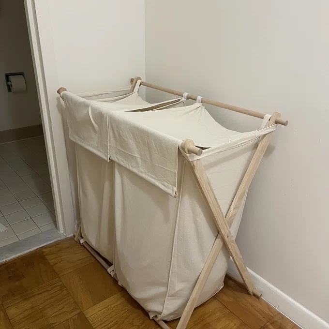 Fabric laundry hamper with wooden frame, placed in a corner on a wooden floor next to a tiled bathroom