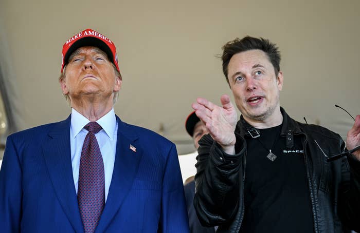 Two well-known public figures are standing together. One wears a red cap and blue suit, the other in a leather jacket gestures while speaking