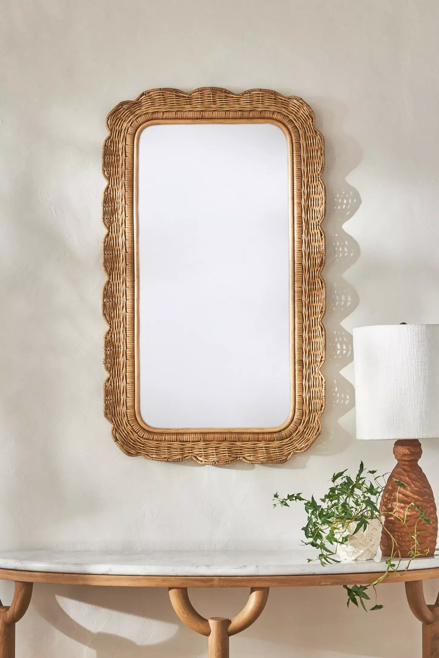 Rectangular rattan mirror with wavy edges on a wall above a marble-topped console with a lamp and greenery