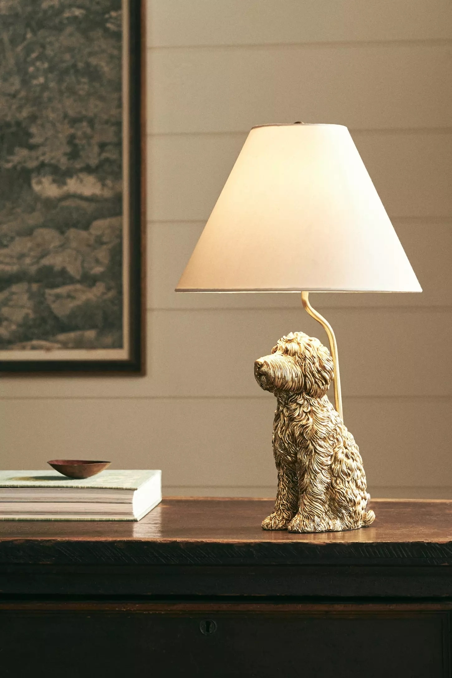 Dog-shaped lamp base with a light-colored shade on a table next to a book and decorative bowl. Cozy interior decor setting