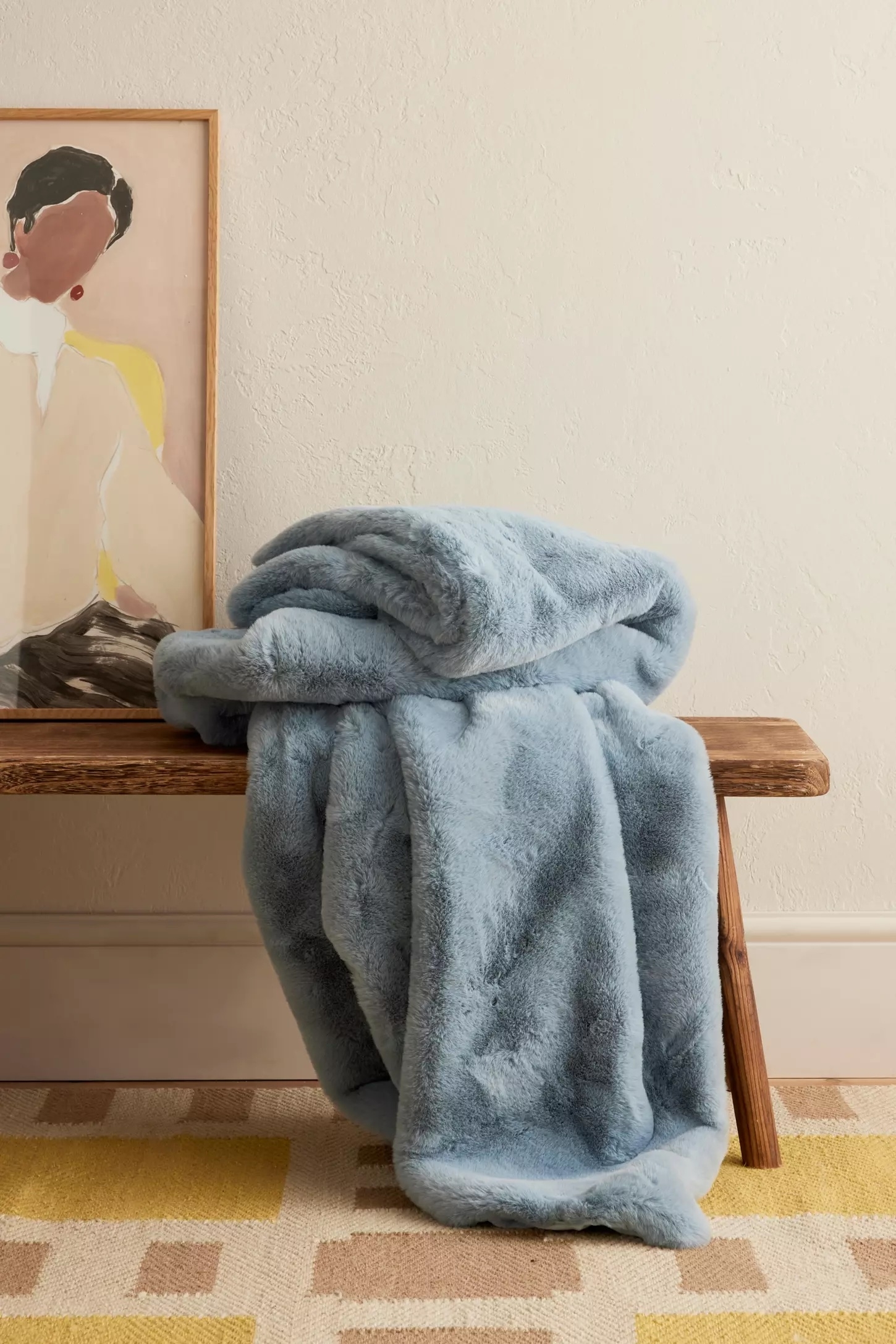 Soft, plush blanket in neutral room, draped on a wooden bench next to a modern art portrait