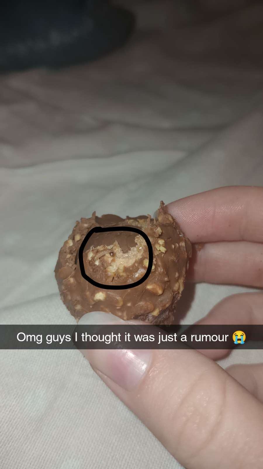 A hand holds a partially eaten Ferrero Rocher chocolate with a visible almond inside. Text overlay: &quot;Omg guys I thought it was just a rumour ?&quot;