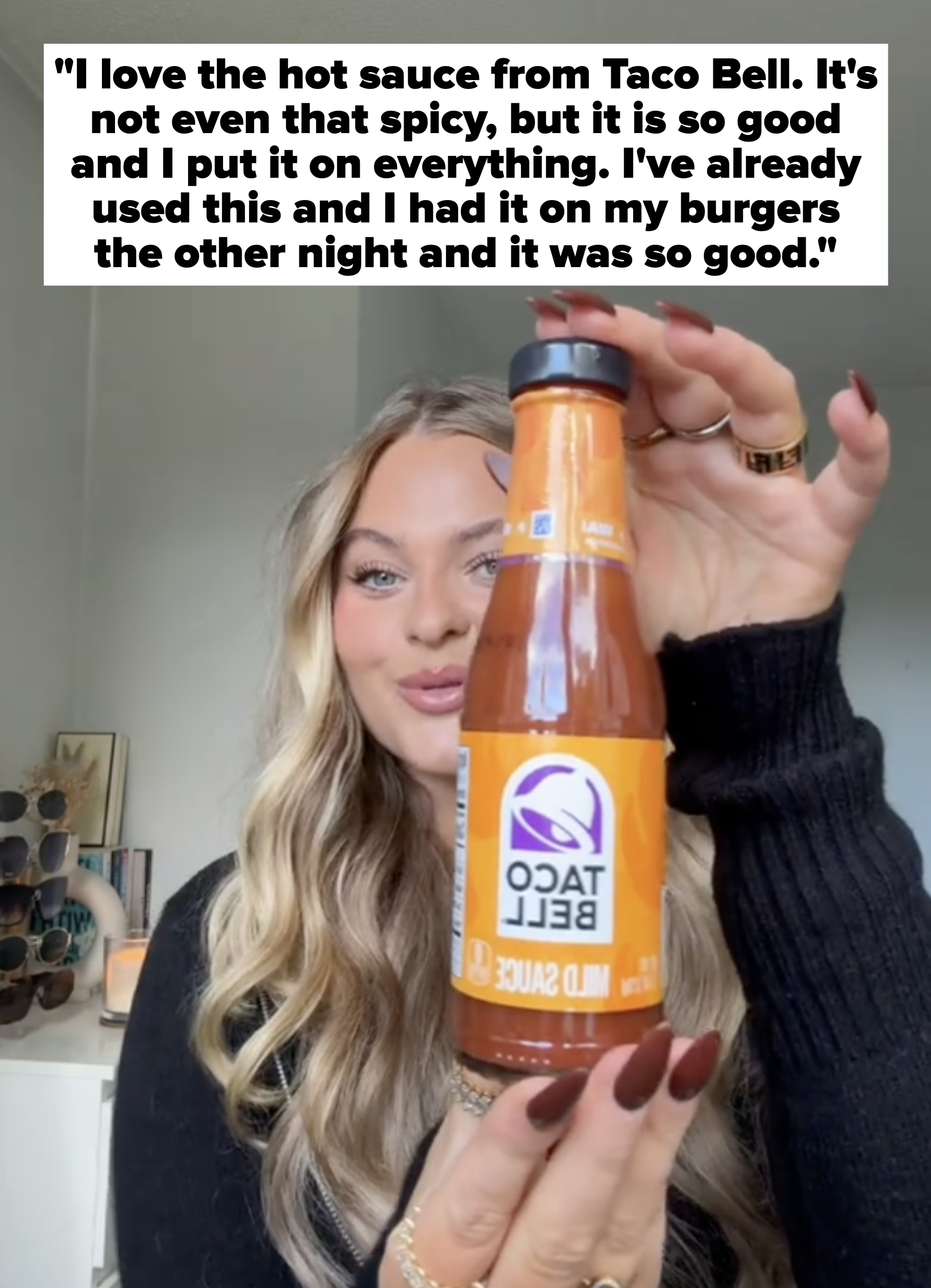 Person holding a bottle of Taco Bell mild sauce, smiling, standing indoors