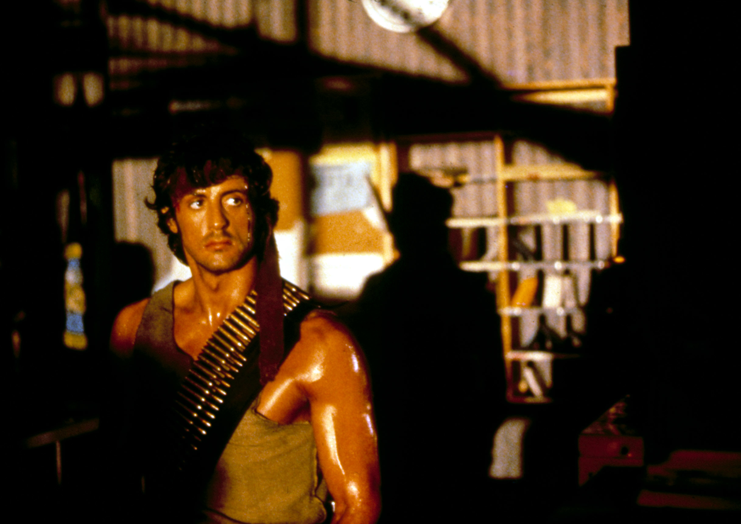 Man in an action scene, wearing a sleeveless top and a bandolier across his chest, with a shadowy background and industrial setting