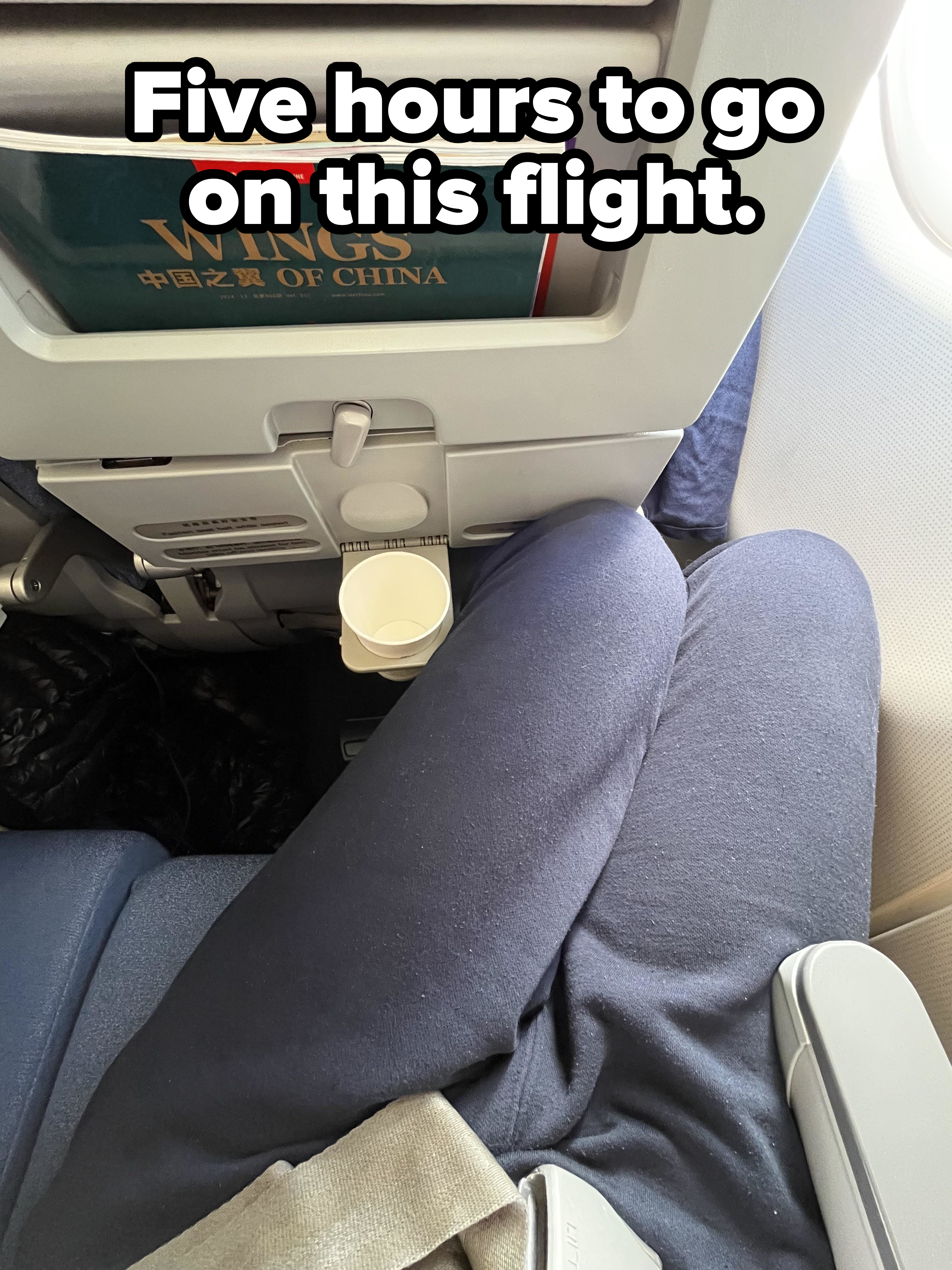 Airplane seat view showing tray table, cup holder, and magazine titled &quot;The Wings of China&quot; in seat pocket