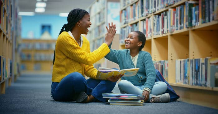 Two people sitting on the floor in a library, smiling and giving a high five, with open books around them