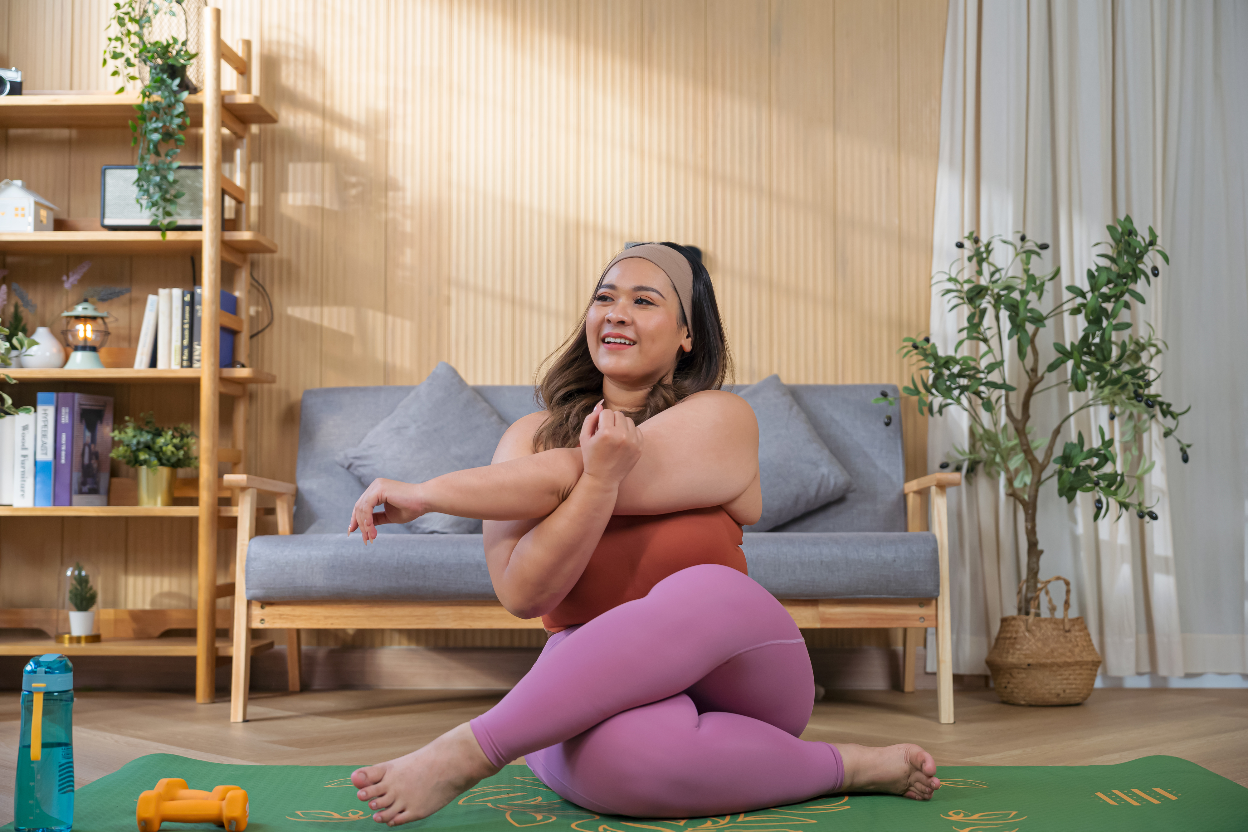 Person sitting cross-legged on a yoga mat, stretching arms, smiling, in a cozy living room with a couch and plants in the background, symbolizing work-life balance