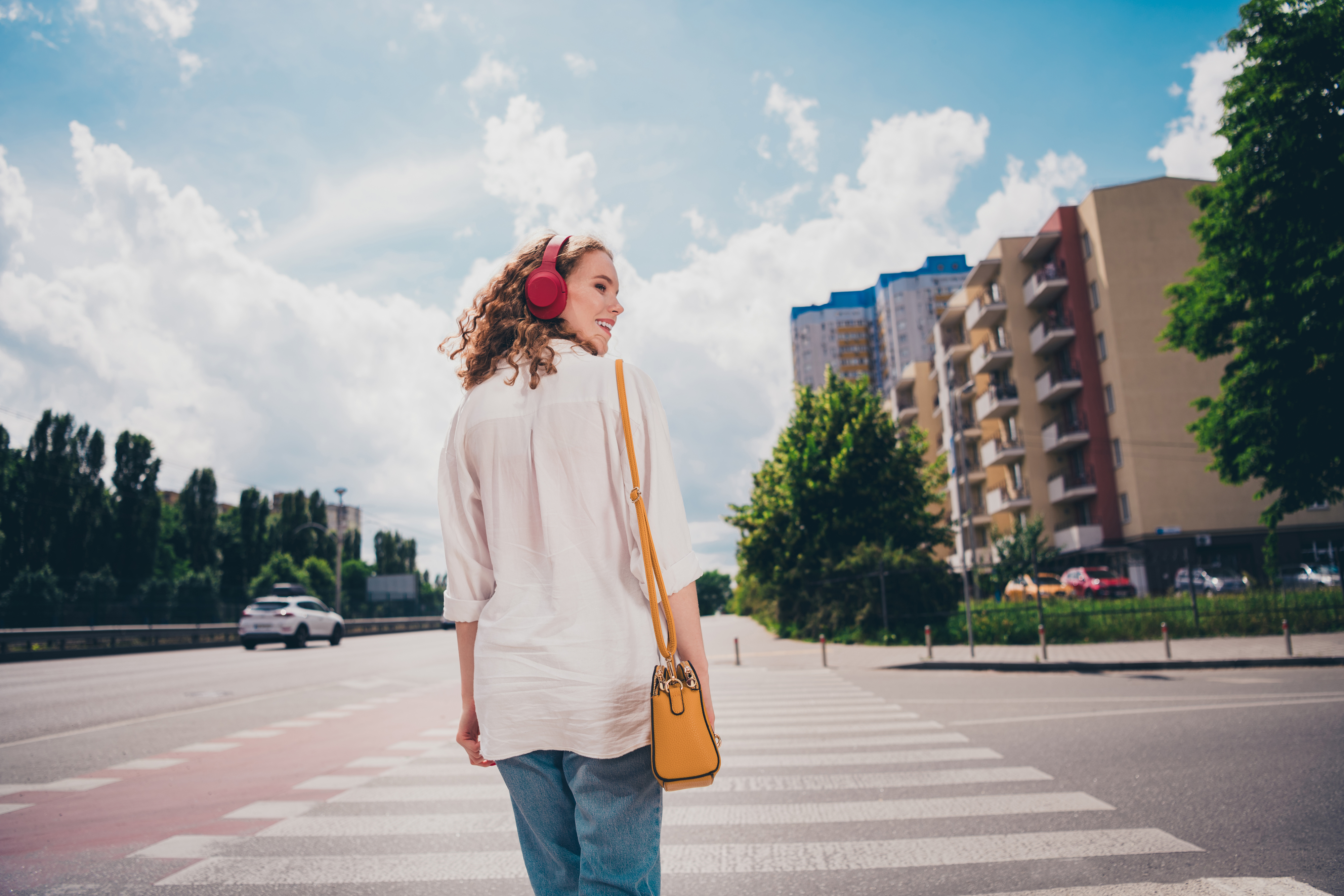 Person wearing headphones and casual outfit walks on a crosswalk, with buildings and trees in the background